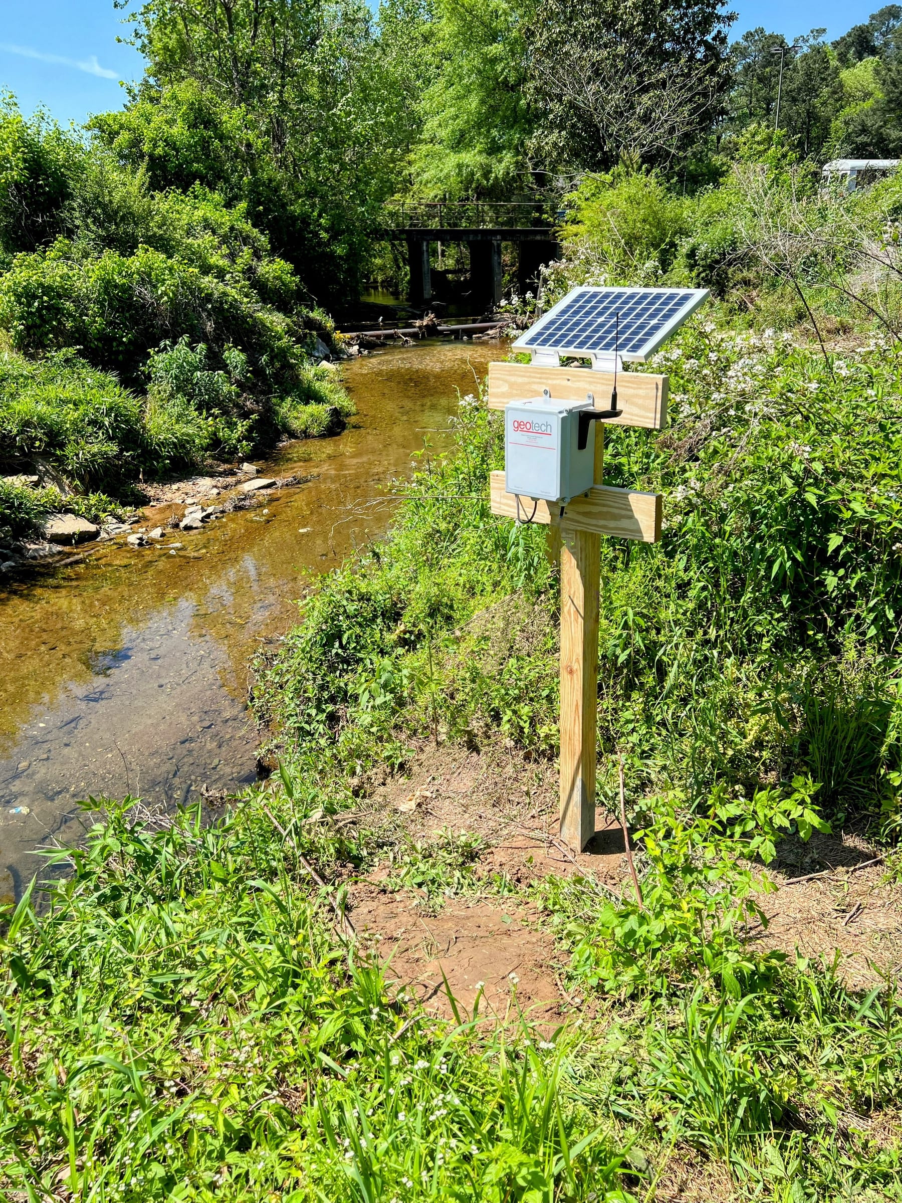 A solar-powered Geotech telemetry station with a pressure sensor mounted on a post beside a shallow, tree-lined creek near Atlanta