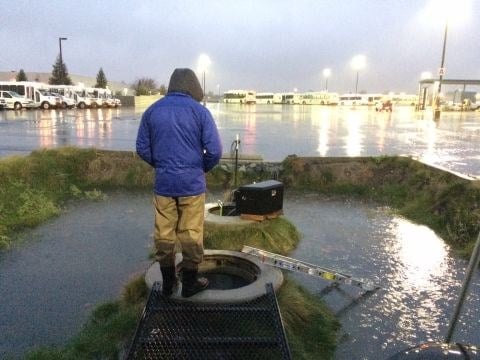 A technician in a rain jacket and waders sampling at a stormwater outfall grate at dusk, with a parking lot of buses in the background