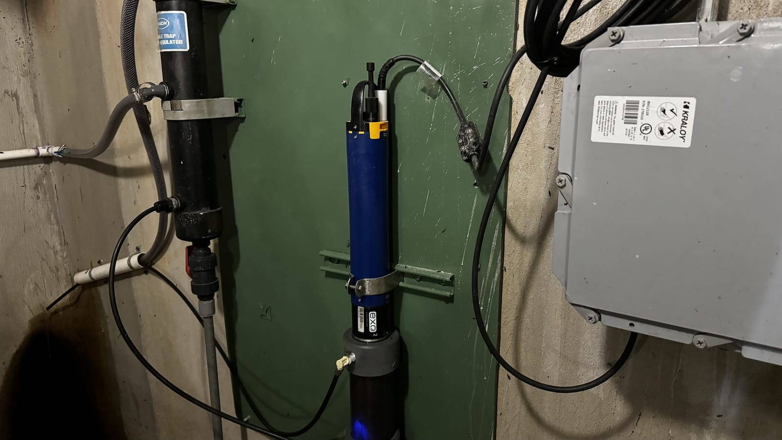 An EXO2 multiparameter water quality sonde mounted in a flow cell on a wall, wired to a control enclosure