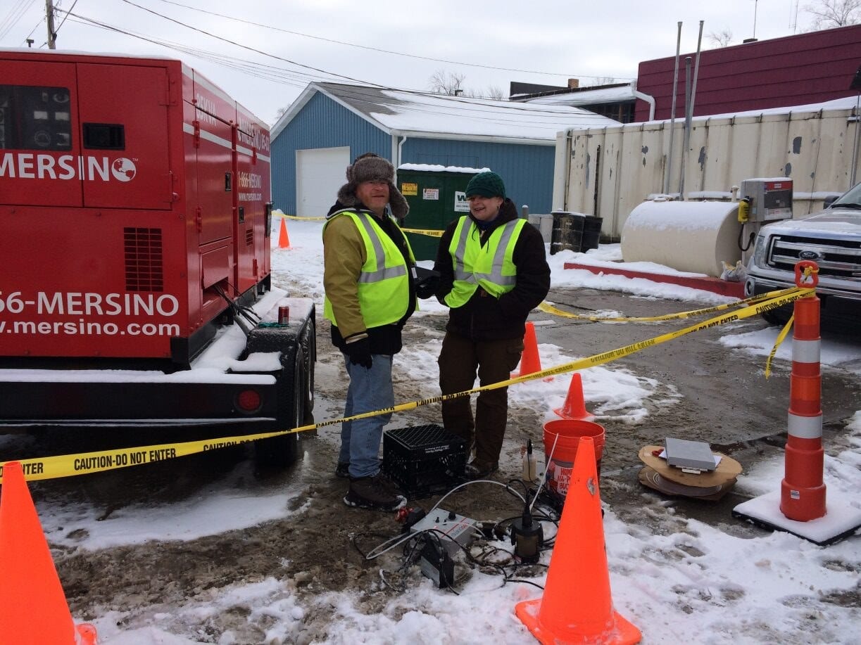 Two technicians in high-visibility vests standing beside a Mersino generator trailer at a cordoned-off winter job site
