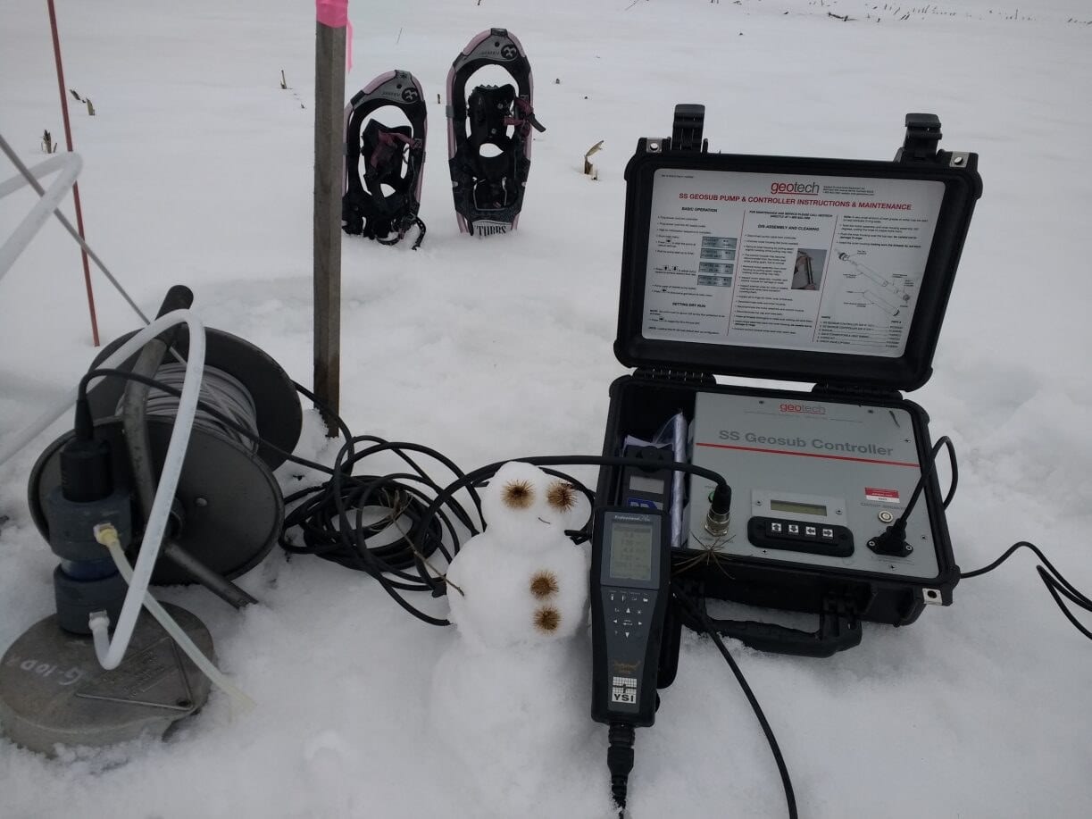 An SS Geosub pump controller case open in the snow next to a cable reel, a handheld YSI ProPlus meter, and a small snowman built beside the equipment