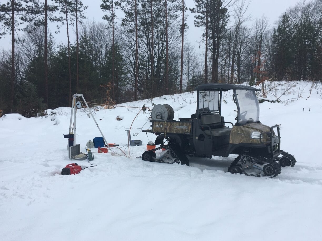 A tracked Kubota utility vehicle and a tripod winch rig set up for well work in a snow-covered forest clearing